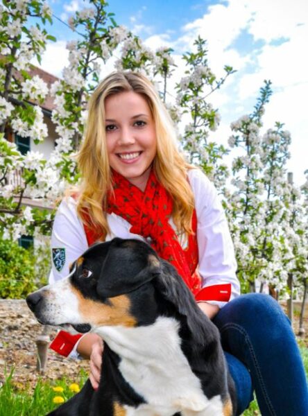 Das Bild zeigt die Obfrau des Reitvereins Ilztal, Bettina Pangerl mit einem schwarz/braun/weißem Hund. Sie selbst hat blonde Haare trägt einen roten Schal mit grünem Muster zu einer weißen Bluse mit Steiermark-Wappen am rechten Ärmel.